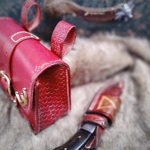Op de afbeelding: Een rood leren tasje met een decoratieve band en een gespsluiting. Het tasje ligt op een wit, pluche oppervlak.