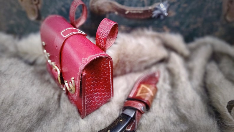 Op de afbeelding: Een rood leren tasje met een decoratieve band en een gespsluiting. Het tasje ligt op een wit, pluche oppervlak.