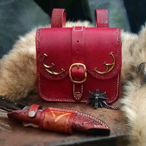 Op de afbeelding: Een rode leren tas met gouden accenten en een gespsluiting. De tas heeft een decoratief ontwerp aan de voorkant en staat op een houten oppervlak. Een klein mes is ook zichtbaar op de afbeelding.