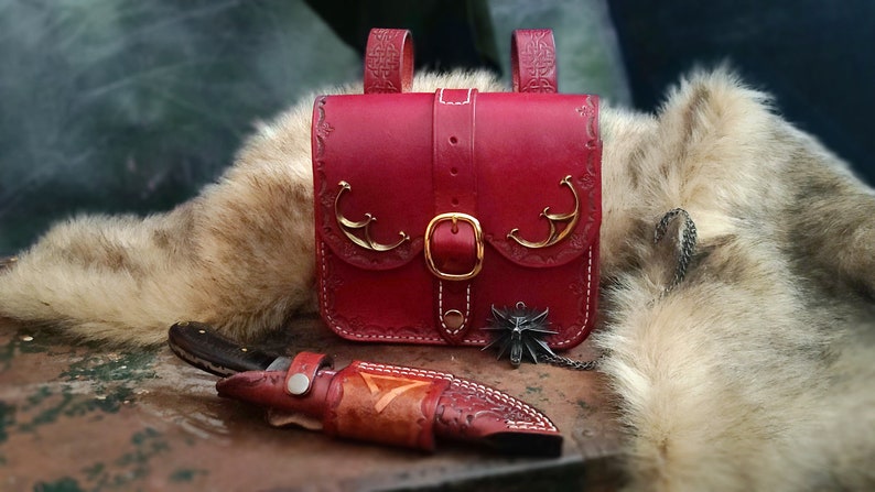Op de afbeelding: Een rode leren tas met gouden accenten en een gespsluiting. De tas heeft een decoratief ontwerp aan de voorkant en staat op een houten oppervlak. Een klein mes is ook zichtbaar op de afbeelding.