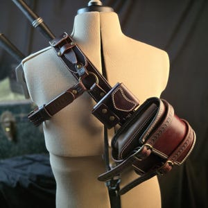 May include: A brown leather shoulder holster with white stitching, featuring multiple straps, a small pouch, and a larger cylindrical holder. The holster is designed to be worn across the chest and around the waist, with brass-colored hardware.