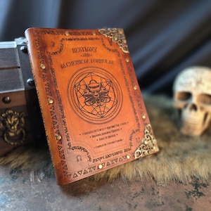 Könnte beinhalten: Ein ledergebundenes Buch mit dem Titel "Bestiarium und Alchemische Formeln" mit aufwendigen goldenen Eckakzenten und einem kreisförmigen Emblem. Das Cover ist braun mit geprägtem Text und einer detaillierten Illustration.