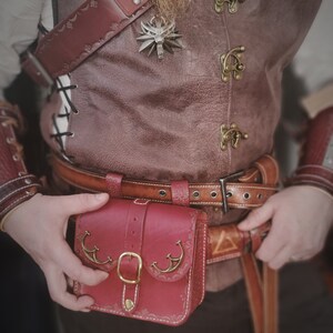 Op de afbeelding: Een rood leren tasje met een decoratieve gesp en metalen accenten. Het tasje is bevestigd aan een bruine leren riem met een zilveren gesp.