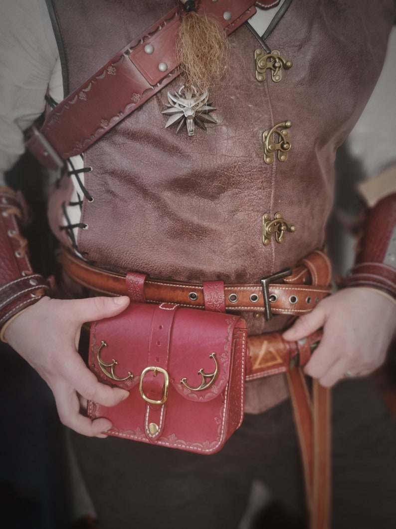Op de afbeelding: Een rood leren tasje met een decoratieve gesp en metalen accenten. Het tasje is bevestigd aan een bruine leren riem met een zilveren gesp.
