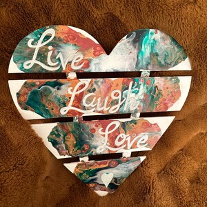 Può includere: Un cartello di legno bianco a forma di cuore con un design di pittura astratta colorata. Il cartello è diviso in quattro sezioni con le parole "Live", "Laugh", "Love" scritte in bianco su ogni sezione.
