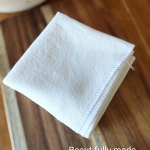 May include: A white cotton dish towel folded in half with a stitched edge. The towel is on a wooden surface.