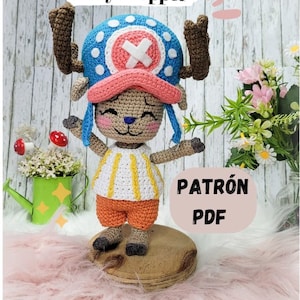 May include: Crocheted amigurumi toy of Tony Chopper from the anime One Piece. The toy is wearing a blue and white hat with a pink brim and a white X on the front. The toy is standing on a wooden base and has a light brown, orange, and white striped shirt. The text "PATRÓN PDF" is visible in the image.