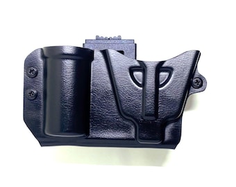 May include: A black, molded polymer holster with a cylindrical magazine holder and a pistol grip compartment. The holster is secured with screws and features a belt clip attachment. The surface has a textured finish.
