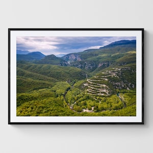 May include: An aerial view of a winding road through a lush green valley. The road is surrounded by trees and mountains, and the sky is cloudy.