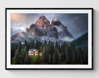 May include: A photograph of a mountain range with a small house in the foreground. The mountain is covered in fog and the sky is a dark blue. The house is lit up with warm light.