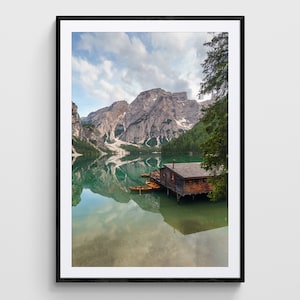 May include: A wooden cabin sits on stilts over a calm, clear lake. The lake is surrounded by mountains and trees. The sky is blue with white clouds.