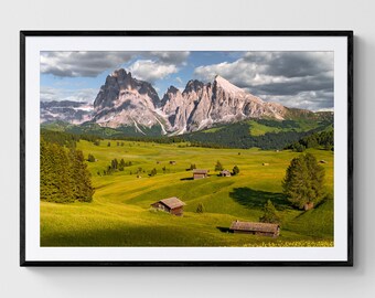 May include: A scenic landscape photograph featuring a mountain range with snow-capped peaks in the background. The foreground shows a lush green meadow with small wooden houses scattered throughout. The sky is a mix of blue and white clouds.