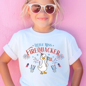 Puede incluir: Camiseta blanca con el texto "LITTLE MISS FIREQUACKER" en rojo y azul. La camiseta presenta un pato de dibujos animados sosteniendo una bandera estadounidense, rodeado de fuegos artificiales y estrellas. Un dise&ntilde;o divertido y festivo para ni&ntilde;os.