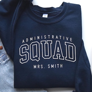 May include: A navy blue sweatshirt with the words "ADMINISTRATIVE SQUAD" and "MRS. SMITH" in white block letters. The sweatshirt is folded on a white surface next to a pair of blue jeans, a brown mug, and a gray sneaker.