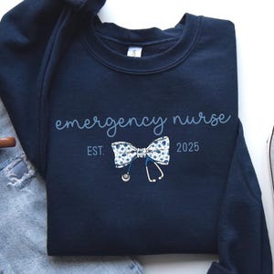 May include: Navy blue sweatshirt with the words "emergency nurse" in script. A bow tie with a floral pattern and a stethoscope graphic are below the text. The year "2025" is printed below the bow tie. A coffee mug, jeans, and a sneaker are in the background.