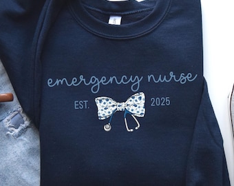 May include: Navy blue sweatshirt with the words "emergency nurse" in script. A bow tie with a floral pattern and a stethoscope graphic are below the text. The year "2025" is printed below the bow tie. A coffee mug, jeans, and a sneaker are in the background.