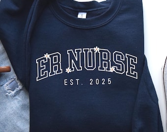May include: A navy blue sweatshirt with the text "ER NURSE" in white block letters, with small flower accents. Below the text is "EST. 2025". The sweatshirt is displayed with a pair of blue jeans, a coffee mug, and a gray sneaker.