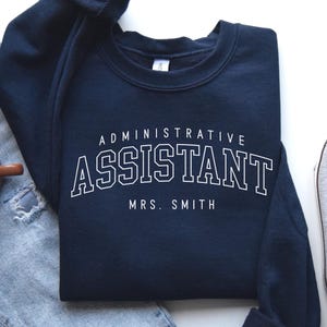 May include: Navy blue sweatshirt with the words "ADMINISTRATIVE ASSISTANT" and "MRS. SMITH" in white block letters. The sweatshirt is folded on top of a pair of blue jeans, a brown mug, and a gray sneaker.