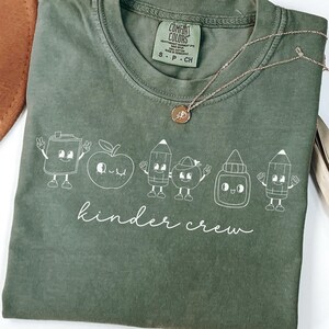 May include: Olive green Comfort Colors t-shirt with a white graphic of cartoon school supplies and the words "kinder crew". A gold necklace with a pendant is draped across the shirt.