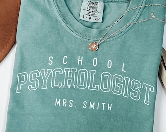 May include: A seafoam green t-shirt with the words "SCHOOL PSYCHOLOGIST MRS. SMITH" in white block letters. A silver necklace with a pendant rests on the shirt. The shirt is folded and has a Comfort Colors tag.