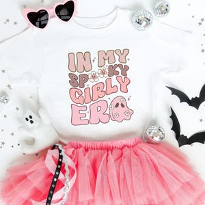 Puede incluir: Una camiseta blanca con la frase "IN MY SPOOKY GIRLY ERA" en letras marrones y rosas, combinada con una falda tut&uacute; rosa. La imagen incluye gafas de sol en forma de coraz&oacute;n, bolas de discoteca y recortes de murci&eacute;lagos, creando un tema de Halloween.