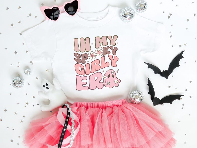 Puede incluir: Una camiseta blanca con la frase "IN MY SPOOKY GIRLY ERA" en letras marrones y rosas, combinada con una falda tut&uacute; rosa. La imagen incluye gafas de sol en forma de coraz&oacute;n, bolas de discoteca y recortes de murci&eacute;lagos, creando un tema de Halloween.