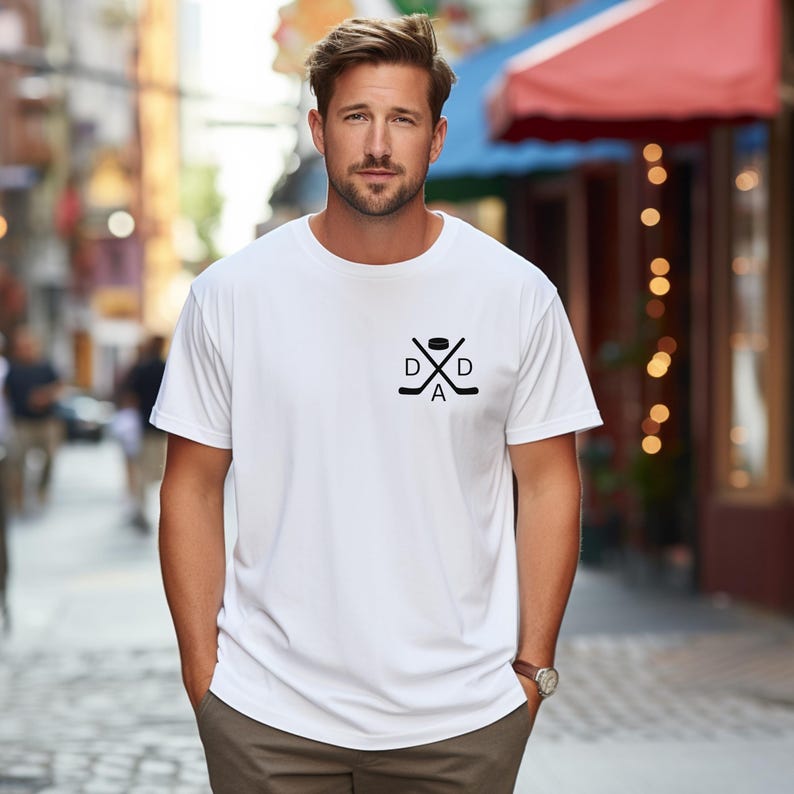 May include: White t-shirt with a black hockey-themed graphic. The graphic includes crossed hockey sticks, a puck, and the word "DAD". The shirt is a crew neck style.