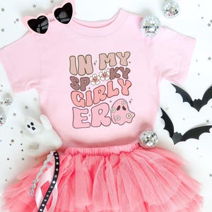 Puede incluir: Una camiseta rosa con el texto "IN MY SPOOKY GIRLY ERA" en letras marrones y rosas, combinada con una falda tut&uacute; rosa y gafas de sol en forma de coraz&oacute;n. El dise&ntilde;o incluye un fantasma.