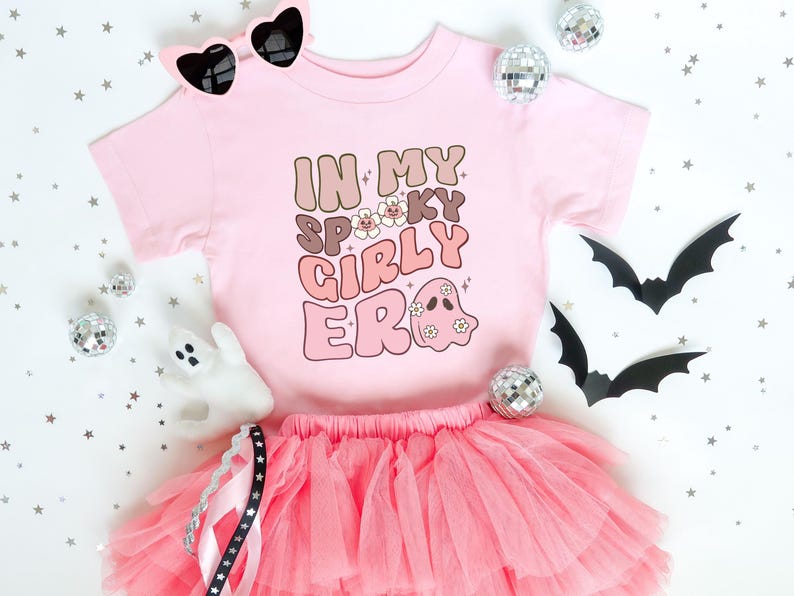 Puede incluir: Una camiseta rosa con el texto "IN MY SPOOKY GIRLY ERA" en letras marrones y rosas, combinada con una falda tut&uacute; rosa y gafas de sol en forma de coraz&oacute;n. El dise&ntilde;o incluye un fantasma.