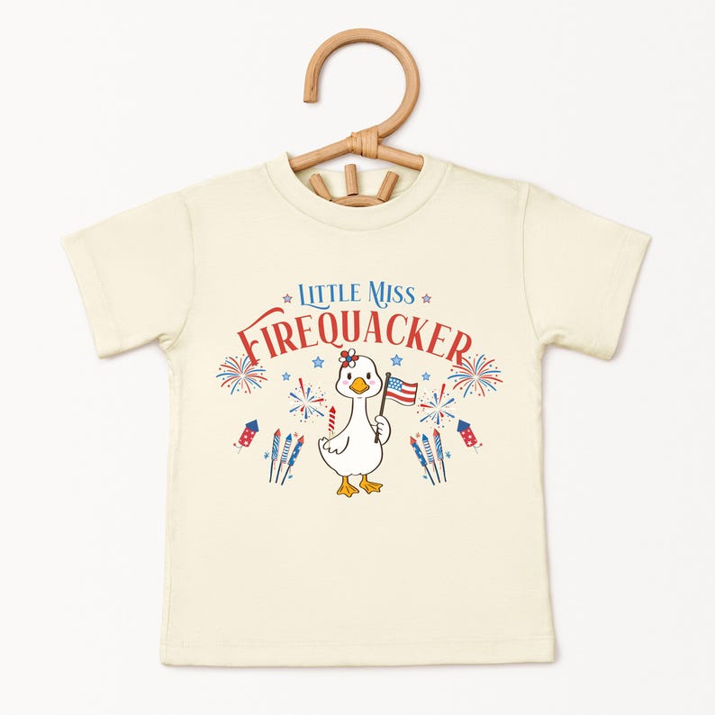 Puede incluir: Camiseta color crema con un pato de dibujos animados sosteniendo una peque&ntilde;a bandera estadounidense. La camiseta tiene el texto "Little Miss Firecracker" en rojo y azul, con gr&aacute;ficos de fuegos artificiales y estrellas. La camiseta est&aacute; colgada en una percha de madera.