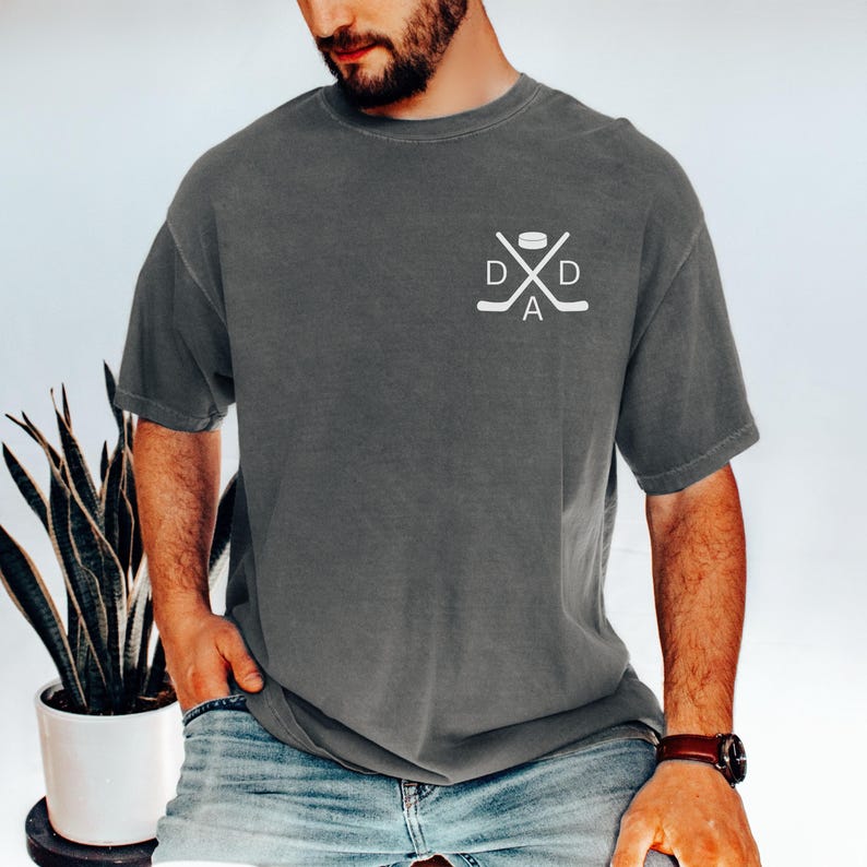 May include: Dark grey t-shirt with a white hockey-themed graphic on the chest. The graphic features crossed hockey sticks, a puck, and the word "DAD". The shirt has short sleeves and a relaxed fit.