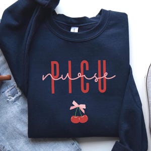 Puede incluir: Sudadera azul marino con el texto "PICU nurse" en rojo y un lazo rosa con dos cerezas rojas debajo.