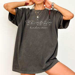 May include: Dark gray t-shirt with a white graphic of school supplies, including an apple, pencils, and glue, with the text "kinder crew" in a script font. The shirt has short sleeves and a relaxed fit.