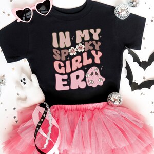 Puede incluir: Camiseta negra con el texto "IN MY SPOOKY GIRLY ERA" en letras rosas y beige, con un gr&aacute;fico de fantasma. Combinada con un tut&uacute; rosa, gafas de sol en forma de coraz&oacute;n y decoraciones de Halloween. Un atuendo divertido y festivo para la temporada de miedo.