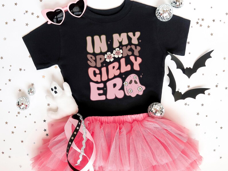 Puede incluir: Camiseta negra con el texto "IN MY SPOOKY GIRLY ERA" en letras rosas y beige, con un gr&aacute;fico de fantasma. Combinada con un tut&uacute; rosa, gafas de sol en forma de coraz&oacute;n y decoraciones de Halloween. Un atuendo divertido y festivo para la temporada de miedo.
