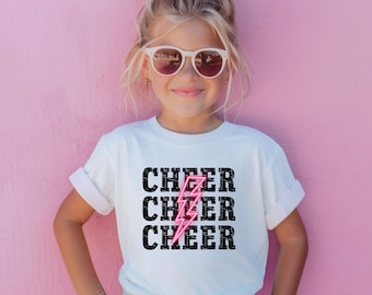 May include: A white t-shirt with the word "CHEER" repeated three times in black text. A pink lightning bolt graphic is centered between the words.