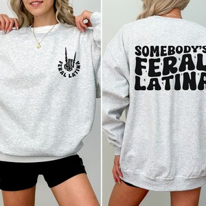 Op de afbeelding: Grijs sweatshirt met een zwarte afbeelding van een hand die een rock-and-roll-teken maakt met de tekst "Feral Latina" op de voorkant. De achterkant van het sweatshirt heeft de tekst "Somebody's Feral Latina" in een retro-lettertype.