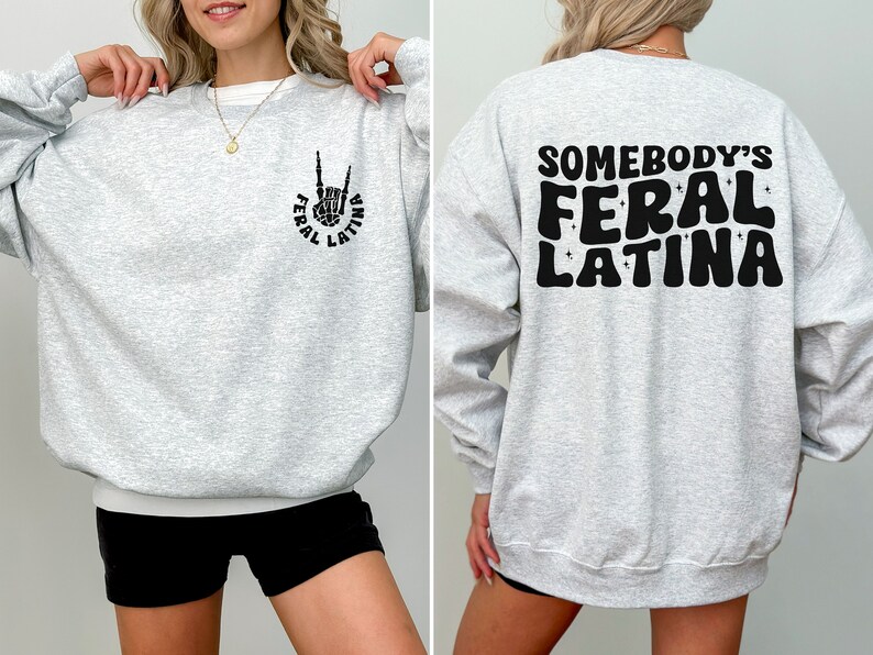 Op de afbeelding: Grijs sweatshirt met een zwarte afbeelding van een hand die een rock-and-roll-teken maakt met de tekst "Feral Latina" op de voorkant. De achterkant van het sweatshirt heeft de tekst "Somebody's Feral Latina" in een retro-lettertype.