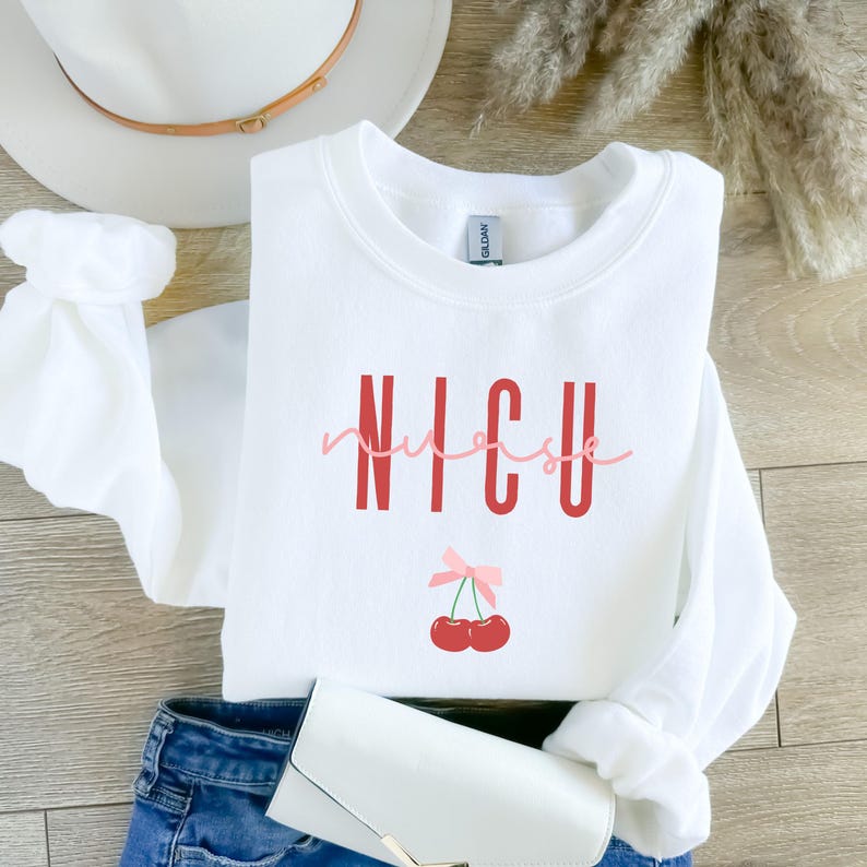 以下が含まれることがあります： 赤い文字で「NICU nurse」と書かれた白いスウェットシャツ。ピンクのリボンと赤いチェリーが2つ下にあります。