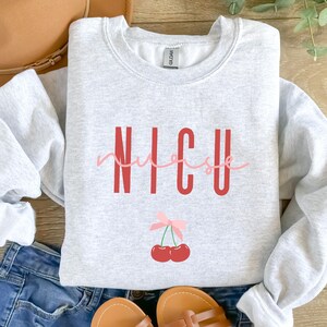 以下が含まれることがあります： 赤い文字で「NICU nurse」と書かれたグレーのスウェットシャツ。テキストの下には、ピンクのリボンと赤いチェリーが2つあります。