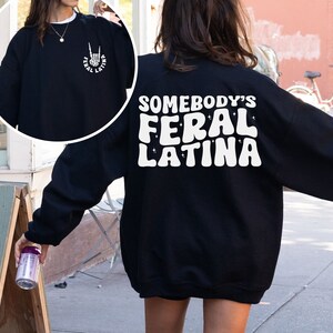 Op de afbeelding: Zwart sweatshirt met witte tekst die "Somebody's Feral Latina" luidt in een retro lettertype. Het sweatshirt heeft een ontspannen pasvorm en lange mouwen.
