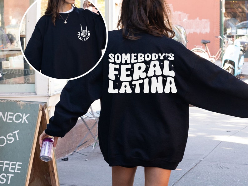 Op de afbeelding: Zwart sweatshirt met witte tekst die "Somebody's Feral Latina" luidt in een retro lettertype. Het sweatshirt heeft een ontspannen pasvorm en lange mouwen.