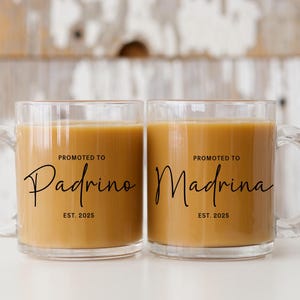 Puede incluir: Dos tazas de café de vidrio transparente con asas negras. Las tazas están llenas de café y tienen el texto "Promoted to Padrino Est. 2025" y "Promoted to Madrina Est. 2025" impreso en ellas.