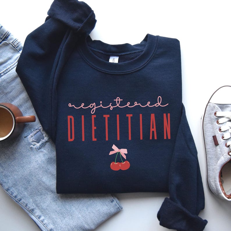 Op de afbeelding: Navyblauwe trui met de tekst "registered dietitian" in rode en roze letters. Onder de tekst bevindt zich een roze strik met twee rode kersen.