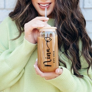 May include: A clear glass tumbler with a bamboo lid and a straw. The tumbler has a floral design and the text "Nina EST. 2024" printed on it.