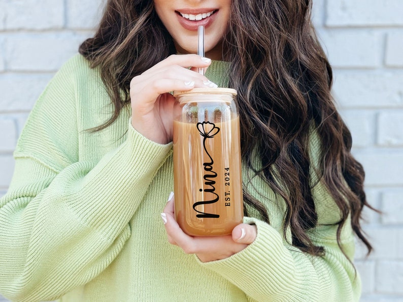 May include: A clear glass tumbler with a bamboo lid and a straw. The tumbler has a floral design and the text "Nina EST. 2024" printed on it.