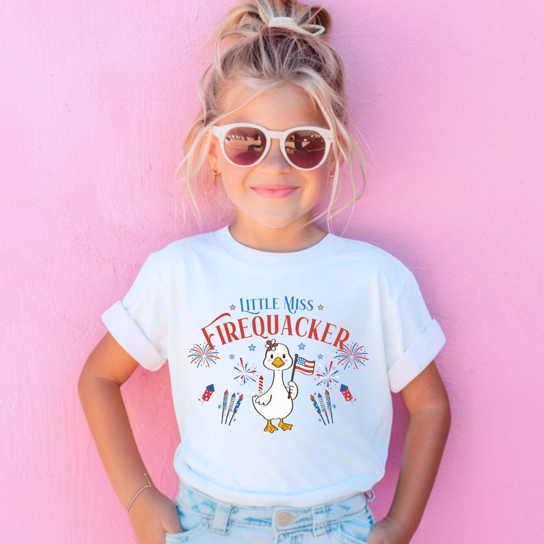 Puede incluir: Camiseta blanca con el texto "LITTLE MISS FIREQUACKER" en rojo y azul. La camiseta presenta un pato de dibujos animados sosteniendo una bandera estadounidense, rodeado de fuegos artificiales y estrellas. Un dise&ntilde;o divertido y festivo para ni&ntilde;os.