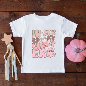 Puede incluir: Camiseta blanca con el texto "IN MY SPOOKY GIRLY ERA" en rosa, marr&oacute;n y beige. Una calabaza rosa y una varita en forma de estrella con cintas est&aacute;n al lado de la camiseta. La camiseta tiene un gr&aacute;fico de fantasma.