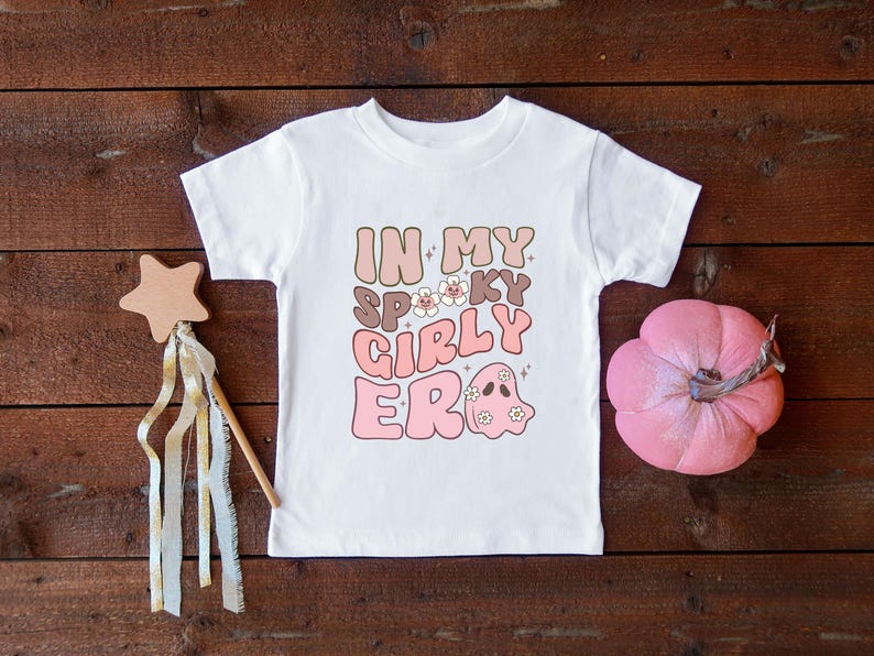 Puede incluir: Camiseta blanca con el texto "IN MY SPOOKY GIRLY ERA" en rosa, marr&oacute;n y beige. Una calabaza rosa y una varita en forma de estrella con cintas est&aacute;n al lado de la camiseta. La camiseta tiene un gr&aacute;fico de fantasma.