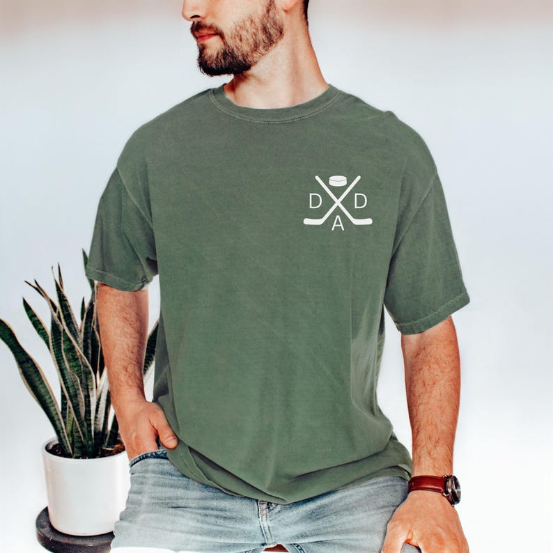 May include: Olive green t-shirt with a white hockey-themed graphic on the chest. The graphic features crossed hockey sticks, a puck, and the word "DAD". The shirt has short sleeves and a relaxed fit.